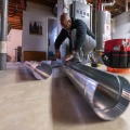 A weatherization installer kneeling on a concrete basement floor adjusts the end of a four inch metal duct section.