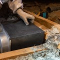 A black exhaust fan is placed between an attic joist cavity by a worker wearing gloves and a white protective coverall. 