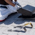 An installer kneeling on a gray asphalt shingled roof holds a black roof vent in place.