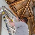 A weatherization installer secures a metal dryer duct to the exterior wall from a ladder.