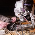 A weatherization installer wearing personal protective equipment kneels on an attic joist while sealing a joint between two pieces of metal ducting using a metallic foil tape. 