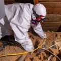 A worker wearing a white protective coverall and respirator moves insulation away from an attic light fixture. 