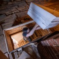 A worker standing on a pull-down stairway ladder holds several pieces of rigid foam over an attic access opening from the attic.
