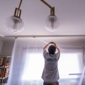 One worker holds a sheet of plastic while the other uses small pieces of tape to hold the plastic to the ceiling of a residential home.