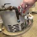A hand turning a red knob on a silver pilot assembly box on a gray water heater. 