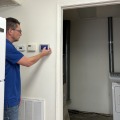 A worker turning off a thermostat by pressing on the digital blue screen of a silver thermostat mounted on a white wall. 